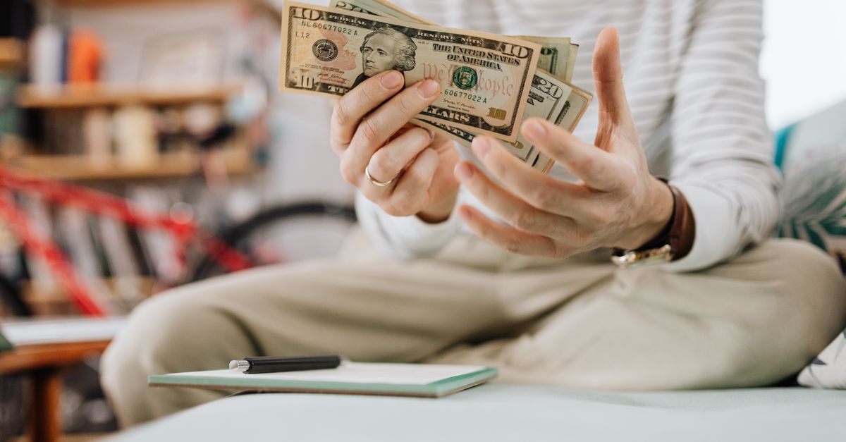 Man wearing casual clothes counts dollar bills while sitting on a sofa indoors.